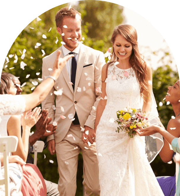 A smiling bride and groom hold hands as they walk down an aisle, while guests throw white flower petals and applaud. The bride holds a bouquet of yellow and white flowers.